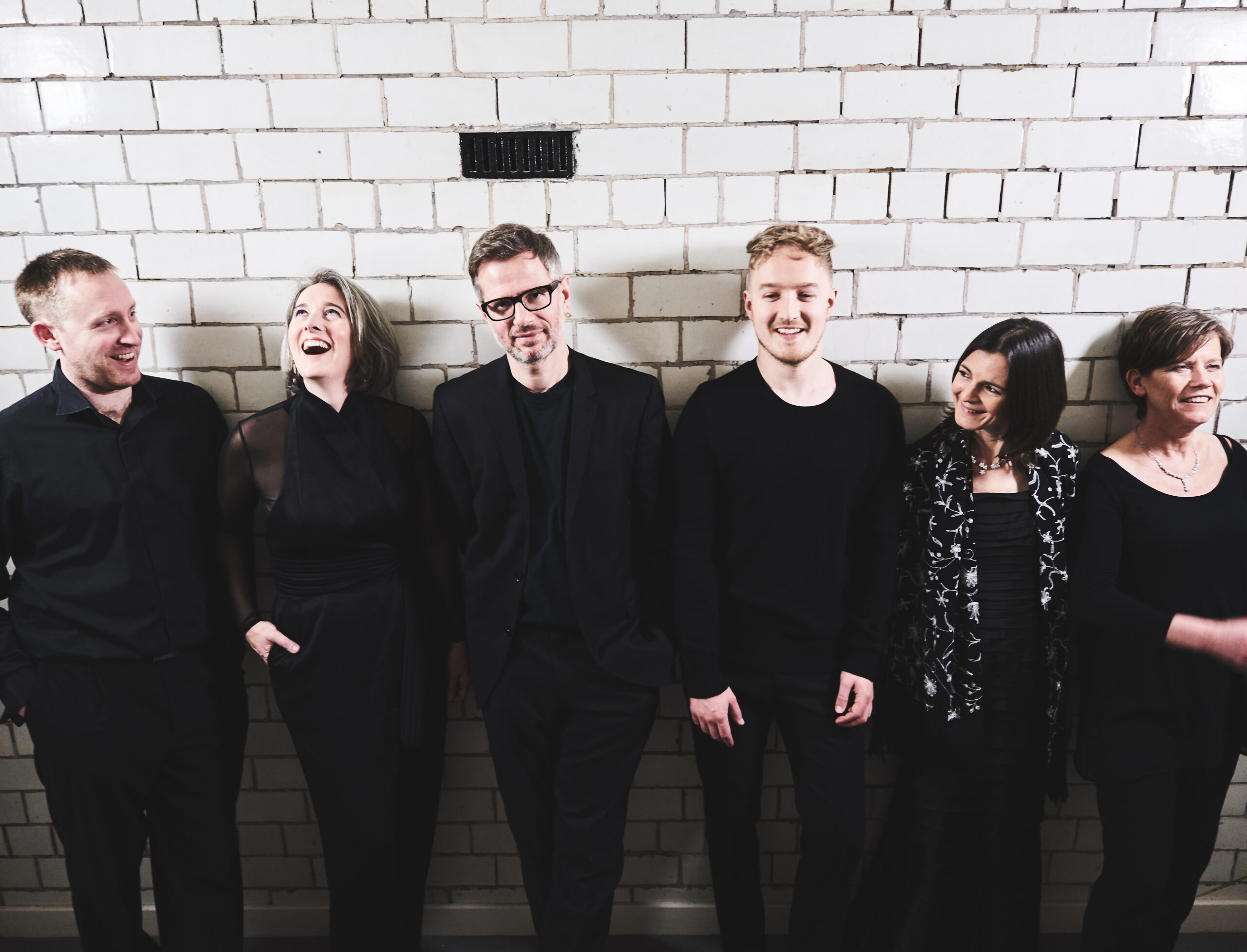 Six ensemble musicians looking relaxed against a faded white brick tiled wall.