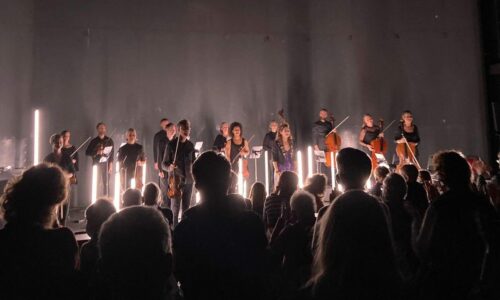 Musicians on stage in front of a concrete wall surrounded by LED light rods and audience sillouettes in foreground.